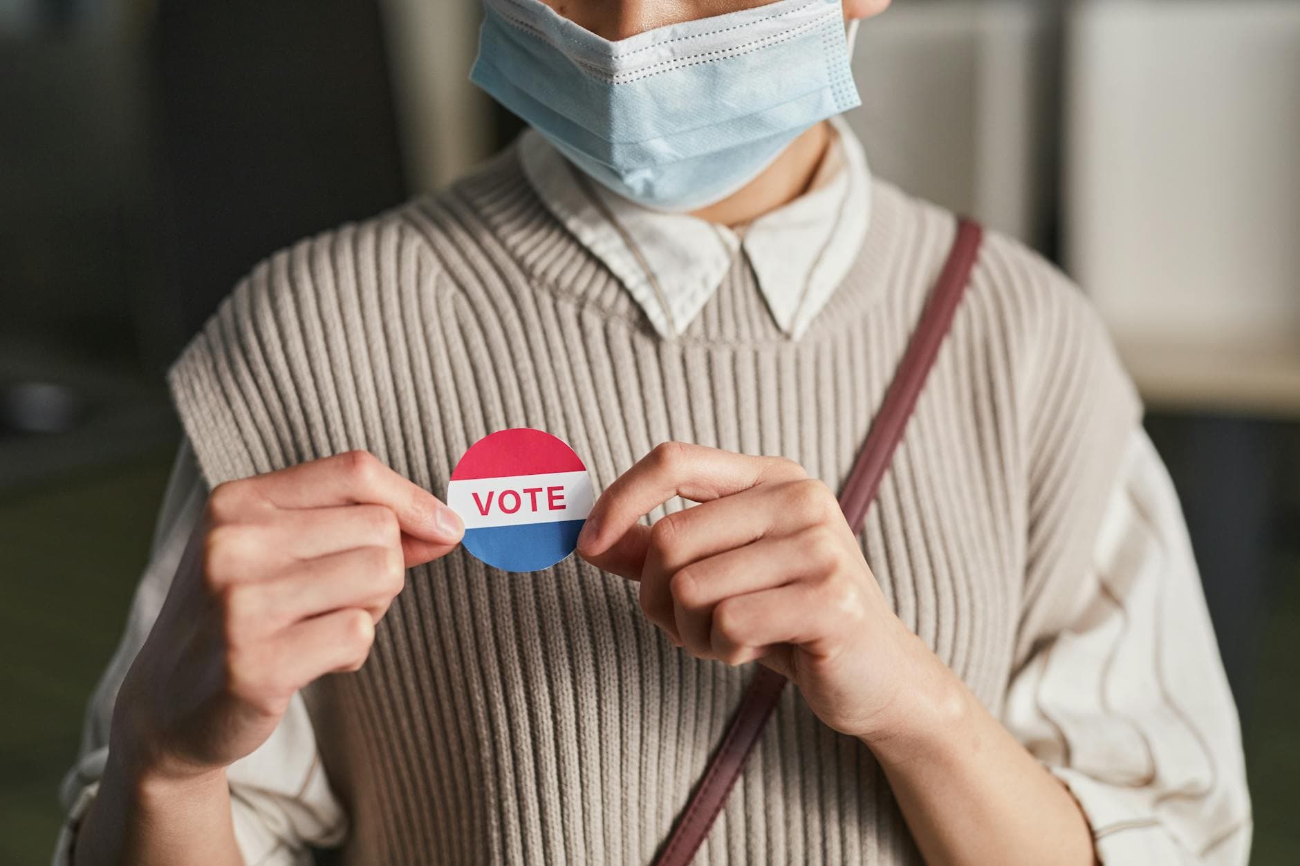 A person wearing a mask holds a voting badge, symbolizing participation in the election.